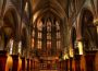 A photo of the inside of a church, a sculpture of crucified Jesus is hanging above the pulpit and behind him is a wall full of stained-glass windows depicting his story.