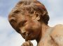 A yellowed stone statue of a child with curly hair holds its finger to its lips in a hushing motion.