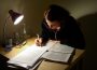 A person at a desk, studying by the light of a desk lamp.
