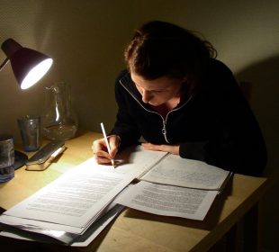 A person at a desk, studying by the light of a desk lamp.