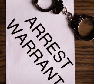 A pair of handcuffs lie on top of a paper that reads “ARREST WARRANT”