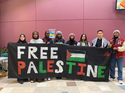 A photo of eight students holding up a sign that says ‘Free Palestine’ in two rows and contains an image of the Palestinian flag beside ‘Free’ and a blood red hand on the top right corner.