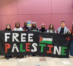 A photo of eight students holding up a sign that says ‘Free Palestine’ in two rows and contains an image of the Palestinian flag beside ‘Free’ and a blood red hand on the top right corner.