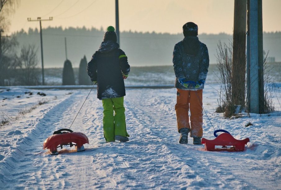 Two children in snow gear pulling red sleds down a snow-covered road.