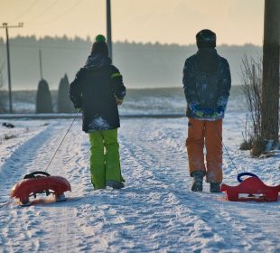 Two children in snow gear pulling red sleds down a snow-covered road.