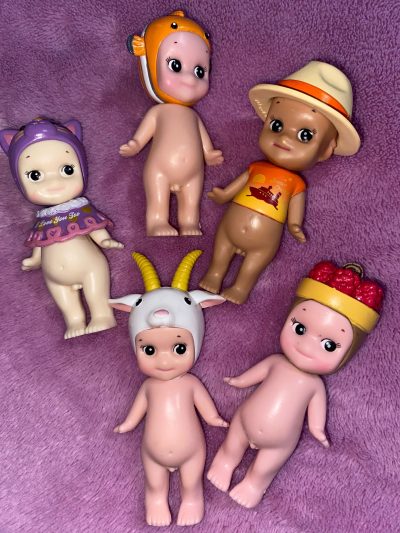 An image of five Sonny Angels on a purple background. There are three on top and two underneath. The top left one is wearing a small purple sweater and a purple cat toque, the bottom left one is wearing a goat toque. The middle top one is wearing a goldfish toque, the top right one is wearing a tan cowboy hat and a sunset orange T-shirt with ‘Sunset Cruise’ as a caption. The bottom right one is wearing a fruit basket on its head.