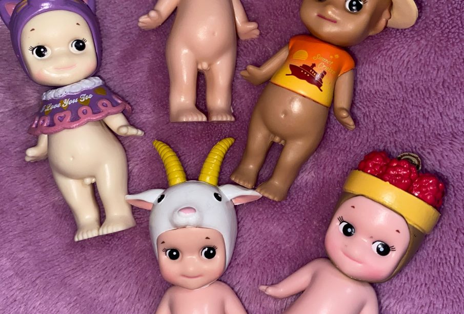 An image of five Sonny Angels on a purple background. There are three on top and two underneath. The top left one is wearing a small purple sweater and a purple cat toque, the bottom left one is wearing a goat toque. The middle top one is wearing a goldfish toque, the top right one is wearing a tan cowboy hat and a sunset orange T-shirt with ‘Sunset Cruise’ as a caption. The bottom right one is wearing a fruit basket on its head.