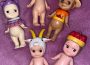 An image of five Sonny Angels on a purple background. There are three on top and two underneath. The top left one is wearing a small purple sweater and a purple cat toque, the bottom left one is wearing a goat toque. The middle top one is wearing a goldfish toque, the top right one is wearing a tan cowboy hat and a sunset orange T-shirt with ‘Sunset Cruise’ as a caption. The bottom right one is wearing a fruit basket on its head.