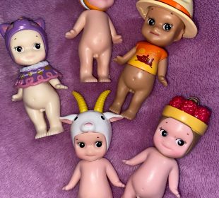 An image of five Sonny Angels on a purple background. There are three on top and two underneath. The top left one is wearing a small purple sweater and a purple cat toque, the bottom left one is wearing a goat toque. The middle top one is wearing a goldfish toque, the top right one is wearing a tan cowboy hat and a sunset orange T-shirt with ‘Sunset Cruise’ as a caption. The bottom right one is wearing a fruit basket on its head.