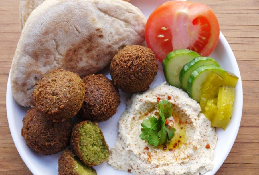An image of a plate filled with two bread pieces called Taboon, four whole falafel and one cut in half, a scoop of hummus topped with cilantro, half of a tomato, three slices of cucumber, and three rectangles of pickles.