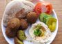 An image of a plate filled with two bread pieces called Taboon, four whole falafel and one cut in half, a scoop of hummus topped with cilantro, half of a tomato, three slices of cucumber, and three rectangles of pickles.