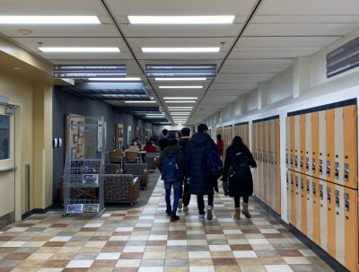 A group of students walk away from the camera through a hallway in the Education building. A Carillon newsstand is in the left corner.