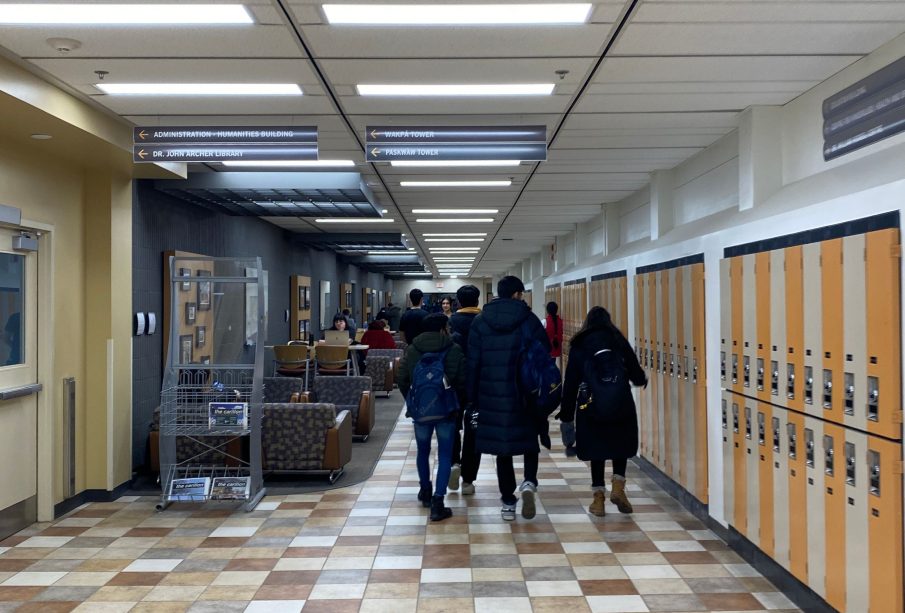 A group of students walk away from the camera through a hallway in the Education building. A Carillon newsstand is in the left corner.