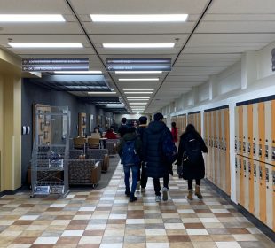 A group of students walk away from the camera through a hallway in the Education building. A Carillon newsstand is in the left corner.