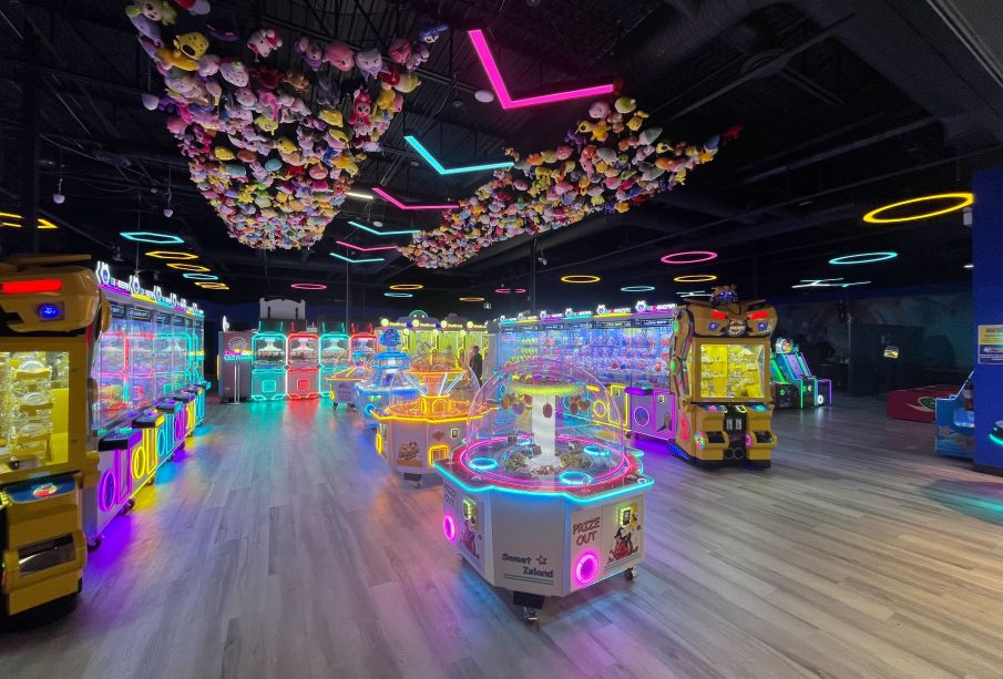 A photo of the VicSquare Arcade, seemingly taken from just inside the entrance and to the left. There are two nets attached to the ceiling full of plushies and various gaming machines and arcade games throughout the image. Only one has a visible name: Sweet Island. There is one person in the background of the image, they are walking out from behind one of the arcade systems.