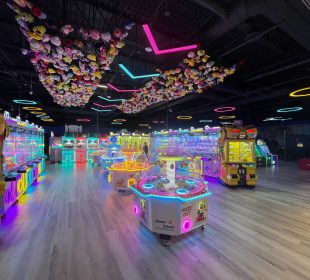 A photo of the VicSquare Arcade, seemingly taken from just inside the entrance and to the left. There are two nets attached to the ceiling full of plushies and various gaming machines and arcade games throughout the image. Only one has a visible name: Sweet Island. There is one person in the background of the image, they are walking out from behind one of the arcade systems.