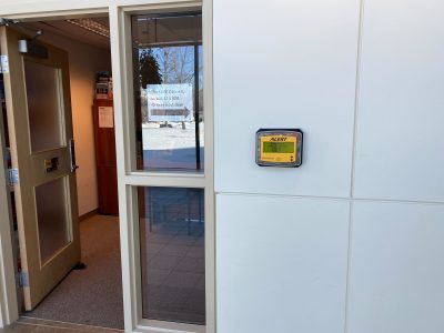 A photo of one of the Alertus wall beacons in the hallway that connects the Paskwāw and Wakpá towers, the Administration and Humanities building, and the CKHS. To the left of the beacon, there is an open door with a printed sign in the window between the door and the beacon. The sign reads “The LINC Office is in WA 114 (ESL Office) next door,” and has an arrow pointing to the right of the photo.