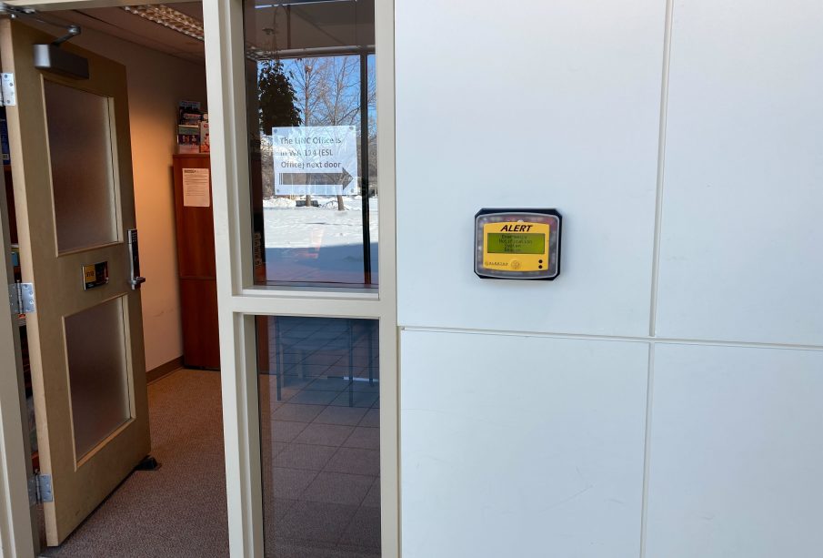 A photo of one of the Alertus wall beacons in the hallway that connects the Paskwāw and Wakpá towers, the Administration and Humanities building, and the CKHS. To the left of the beacon, there is an open door with a printed sign in the window between the door and the beacon. The sign reads “The LINC Office is in WA 114 (ESL Office) next door,” and has an arrow pointing to the right of the photo.