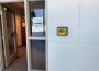 A photo of one of the Alertus wall beacons in the hallway that connects the Paskwāw and Wakpá towers, the Administration and Humanities building, and the CKHS. To the left of the beacon, there is an open door with a printed sign in the window between the door and the beacon. The sign reads “The LINC Office is in WA 114 (ESL Office) next door,” and has an arrow pointing to the right of the photo.