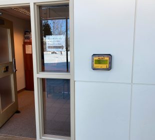 A photo of one of the Alertus wall beacons in the hallway that connects the Paskwāw and Wakpá towers, the Administration and Humanities building, and the CKHS. To the left of the beacon, there is an open door with a printed sign in the window between the door and the beacon. The sign reads “The LINC Office is in WA 114 (ESL Office) next door,” and has an arrow pointing to the right of the photo.