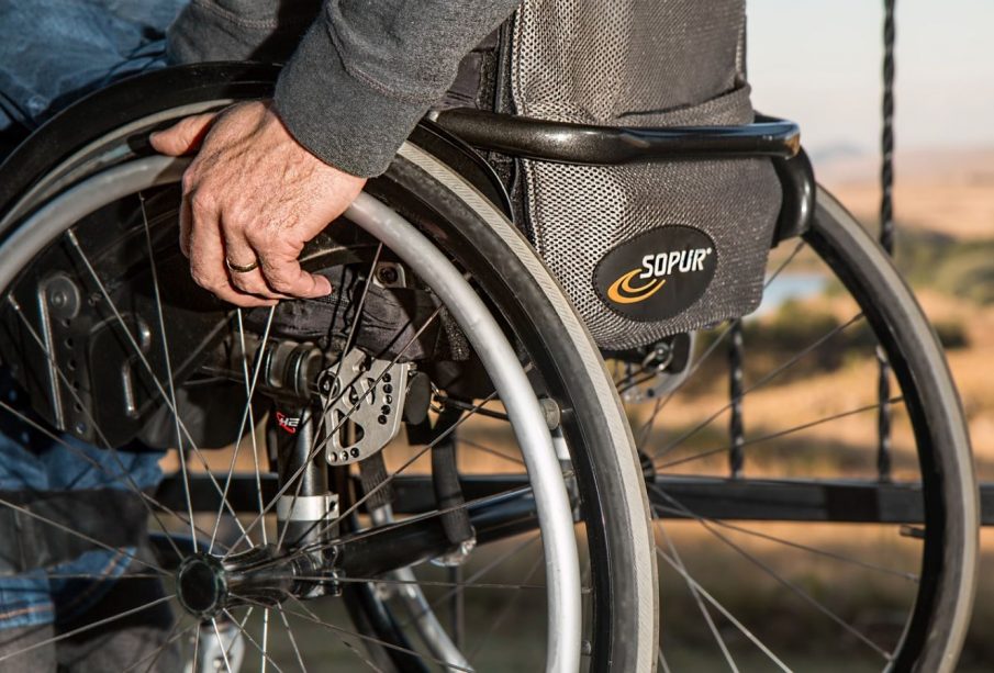A person’s hand rests on their wheelchair, they are outdoors and wearing jeans and a sweatshirt.