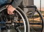 A person’s hand rests on their wheelchair, they are outdoors and wearing jeans and a sweatshirt.