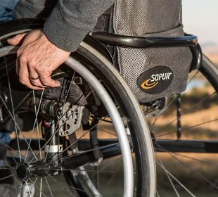 A person’s hand rests on their wheelchair, they are outdoors and wearing jeans and a sweatshirt.