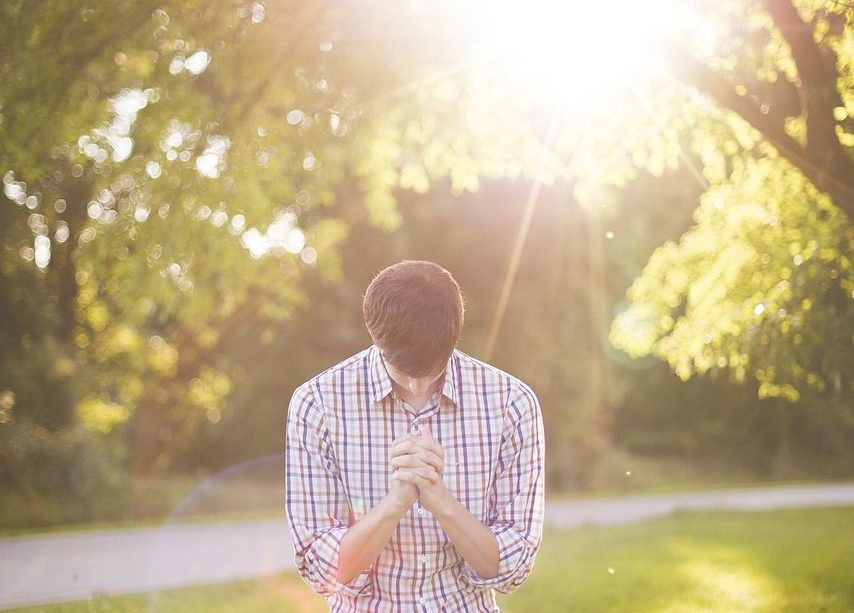 A person on their knees on the grass in summer with a tree behind them on their right. They are holding their hands and bowing their head in prayer while the sun shines behind them and results in a lens flare to the left of them.