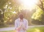 A person on their knees on the grass in summer with a tree behind them on their right. They are holding their hands and bowing their head in prayer while the sun shines behind them and results in a lens flare to the left of them.