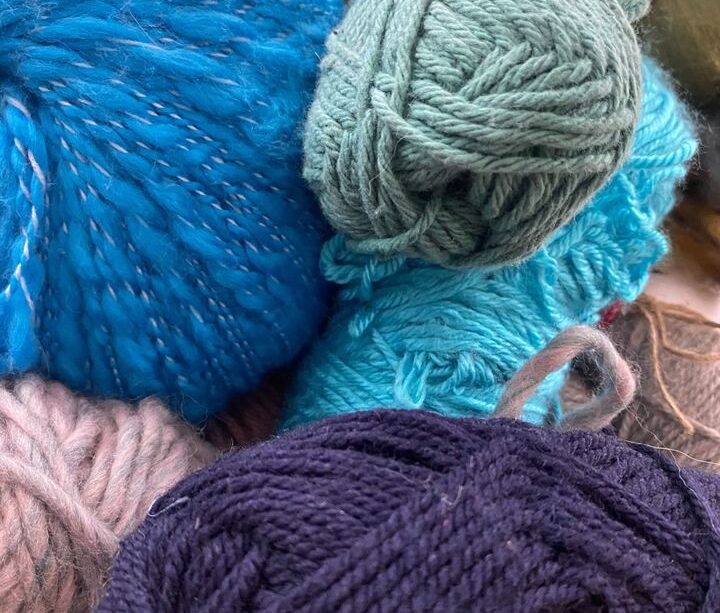 A photograph of the author’s knitting supplies. There are multiple sets and colours of yarn. The ones in-focus are blue, purple, pink, and a muted green. In the top left corner is a multi-coloured project with a knitting needle sticking out of it.