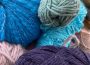 A photograph of the author’s knitting supplies. There are multiple sets and colours of yarn. The ones in-focus are blue, purple, pink, and a muted green. In the top left corner is a multi-coloured project with a knitting needle sticking out of it.