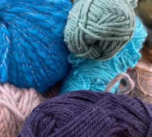 A photograph of the author’s knitting supplies. There are multiple sets and colours of yarn. The ones in-focus are blue, purple, pink, and a muted green. In the top left corner is a multi-coloured project with a knitting needle sticking out of it.
