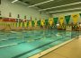 A photo of the Aquatic Centre’s lap swimming pool while empty. The pool is separated into five 25 metre swimming lanes by four yellow and green dividers, and two yellow and green Cougars swimming banners hang across the width of the pool. There are flags hanging on the wall and photos of people above them. Along the back wall is a shelf full of lifejackets, a whiteboard with writing on it and some desks.