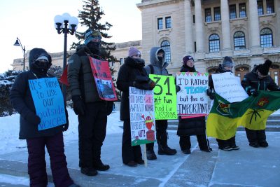Seven Saskatchewan teachers stand in front of the Saskatchewan parliament building. They are dressed in winter clothing and holding signs. One bright green sign reads “-31 isn’t fun //fund our cla$$rooms.”