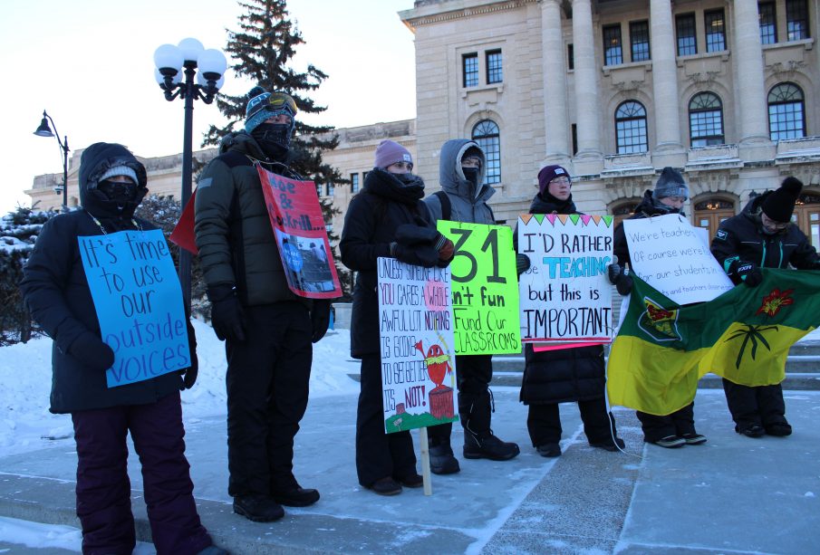 Seven Saskatchewan teachers stand in front of the Saskatchewan parliament building. They are dressed in winter clothing and holding signs. One bright green sign reads “-31 isn’t fun //fund our cla$$rooms.”
