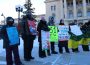 Seven Saskatchewan teachers stand in front of the Saskatchewan parliament building. They are dressed in winter clothing and holding signs. One bright green sign reads “-31 isn’t fun //fund our cla$$rooms.”