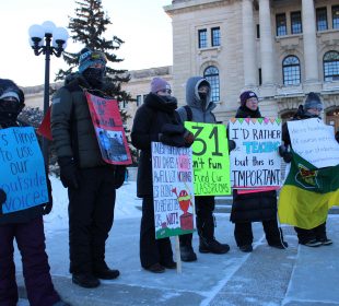 Seven Saskatchewan teachers stand in front of the Saskatchewan parliament building. They are dressed in winter clothing and holding signs. One bright green sign reads “-31 isn’t fun //fund our cla$$rooms.”