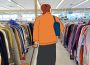 A drawing of a person pulling a shopping cart full of clothes overlaid over a photograph of an aisle of sweaters or cardigans in a thrift store.
