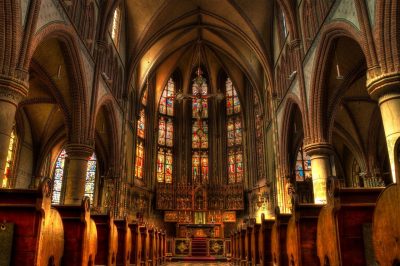 A photo of the inside of a church, a sculpture of crucified Jesus is hanging above the pulpit and behind him is a wall full of stained-glass windows depicting his story.