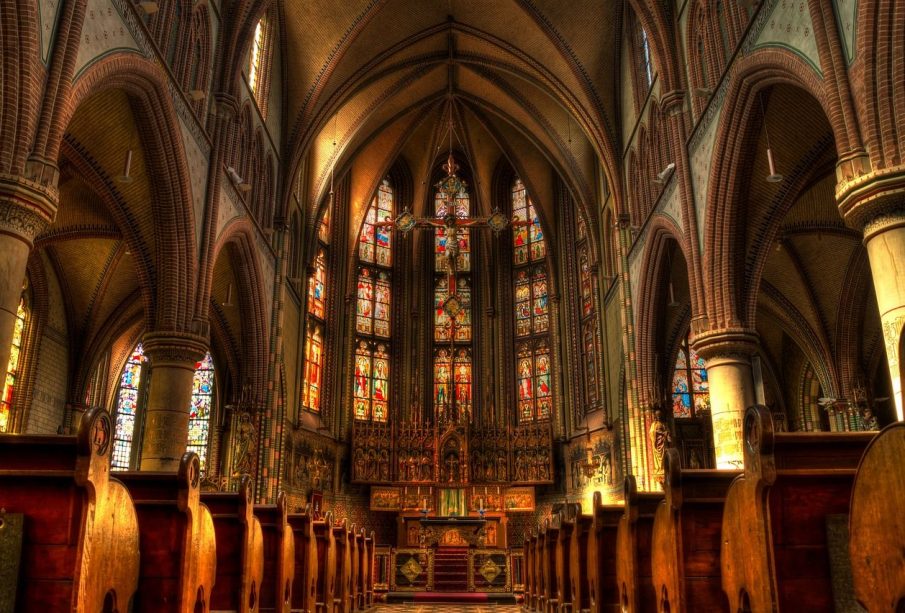 A photo of the inside of a church, a sculpture of crucified Jesus is hanging above the pulpit and behind him is a wall full of stained-glass windows depicting his story.