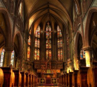 A photo of the inside of a church, a sculpture of crucified Jesus is hanging above the pulpit and behind him is a wall full of stained-glass windows depicting his story.