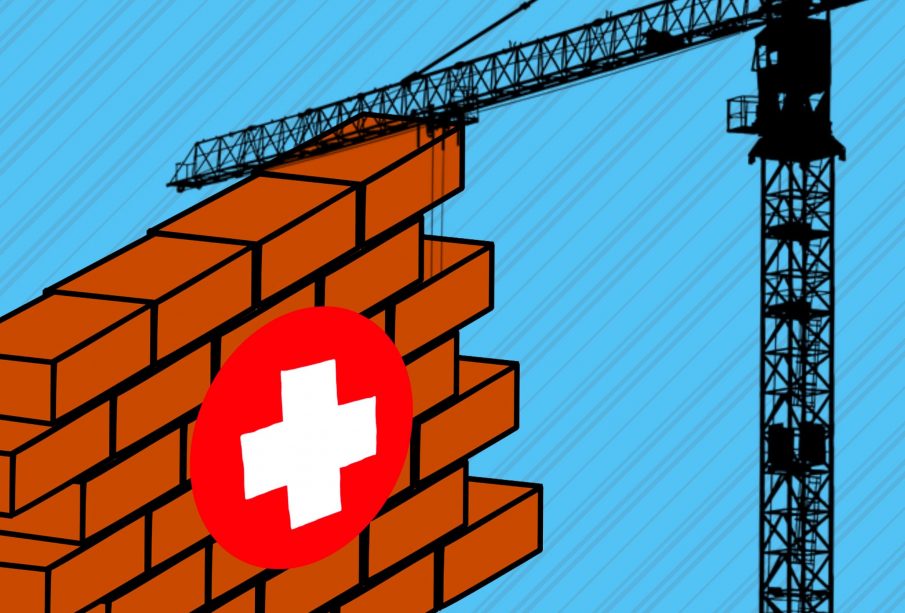 A section of a brick wall with a first aid sign on its front. A solid black silhouette of a crane is lifting the brick wall.
