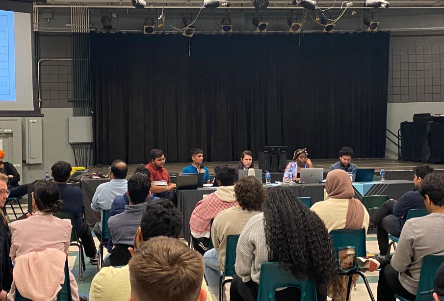 Students sit in attendance at URSU’s AGM. Four rows full of students are seen seated directly in front of the board. 