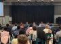 Students sit in attendance at URSU’s AGM. Four rows full of students are seen seated directly in front of the board.