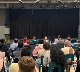 Students sit in attendance at URSU’s AGM. Four rows full of students are seen seated directly in front of the board. 
