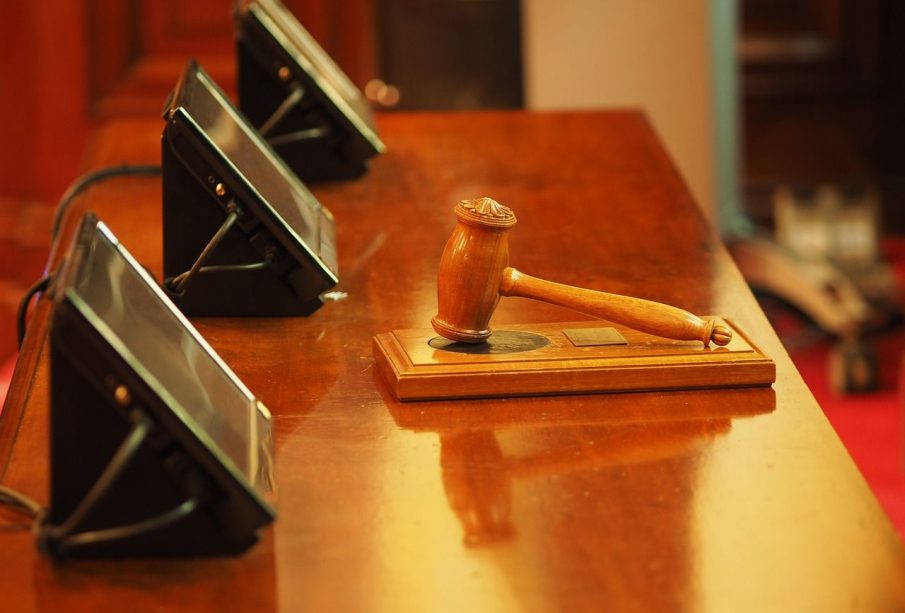 A photo of a gavel resting on a sound block on a wooden desk in front of three rectangular screens.
