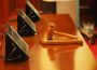 A photo of a gavel resting on a sound block on a wooden desk in front of three rectangular screens.