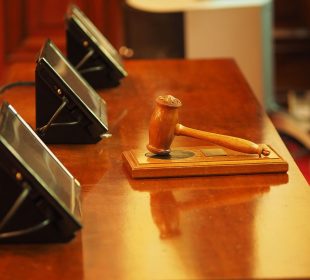 A photo of a gavel resting on a sound block on a wooden desk in front of three rectangular screens.