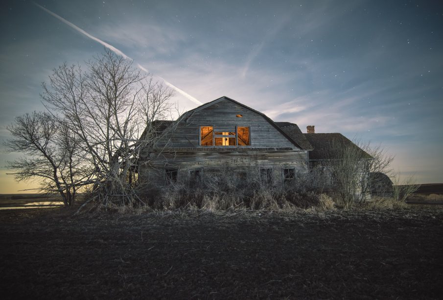 A photograph of the eastern side of an old and sun-faded wooden house. The windows are missing their panes and the wood is gray with age. There are leafless trees, seemingly because of it being early spring or late winter when the photo was taken, along the edge of the building and a light seems to be glowing from inside the house. The sun is setting on the other side of the building and some stars are twinkling in the lightly clouded blue sky above the building. 