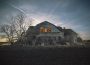 A photograph of the eastern side of an old and sun-faded wooden house. The windows are missing their panes and the wood is gray with age. There are leafless trees, seemingly because of it being early spring or late winter when the photo was taken, along the edge of the building and a light seems to be glowing from inside the house. The sun is setting on the other side of the building and some stars are twinkling in the lightly clouded blue sky above the building. 